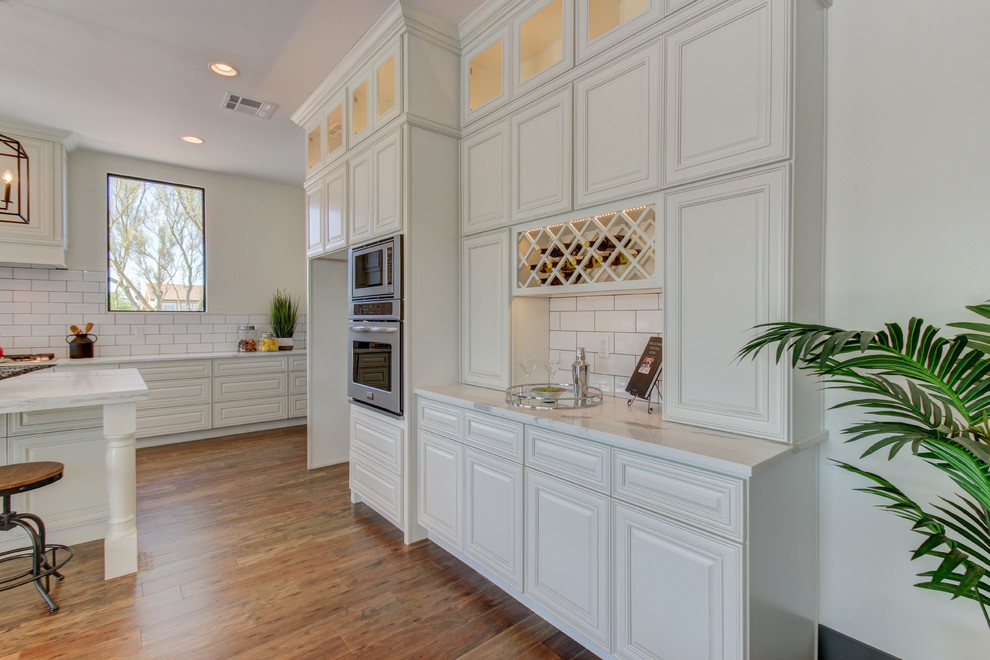 Inspiration for a mid-sized transitional l-shaped dark wood floor and brown floor eat-in kitchen remodel in Phoenix with a farmhouse sink, raised-panel cabinets, white cabinets, marble countertops, white backsplash, subway tile backsplash, stainless steel appliances and an island