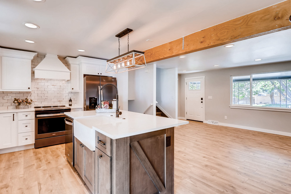 Photo of a large country l-shaped open plan kitchen in Denver with a farmhouse sink, shaker cabinets, white cabinets, quartz benchtops, white splashback, ceramic splashback, stainless steel appliances, laminate floors, with island, brown floor and white benchtop.