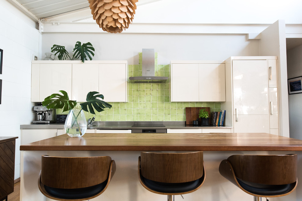 Mid-sized industrial single-wall eat-in kitchen in London with an integrated sink, flat-panel cabinets, white cabinets, stainless steel benchtops, green splashback, ceramic splashback, stainless steel appliances, laminate floors, with island, brown floor and grey benchtop.