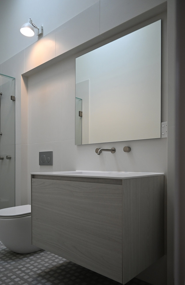 Bild på ett litet funkis vit vitt badrum för barn, med släta luckor, vita skåp, en dusch i en alkov, en vägghängd toalettstol, vit kakel, keramikplattor, vita väggar, klinkergolv i keramik, ett väggmonterat handfat, bänkskiva i akrylsten, grått golv och dusch med gångjärnsdörr