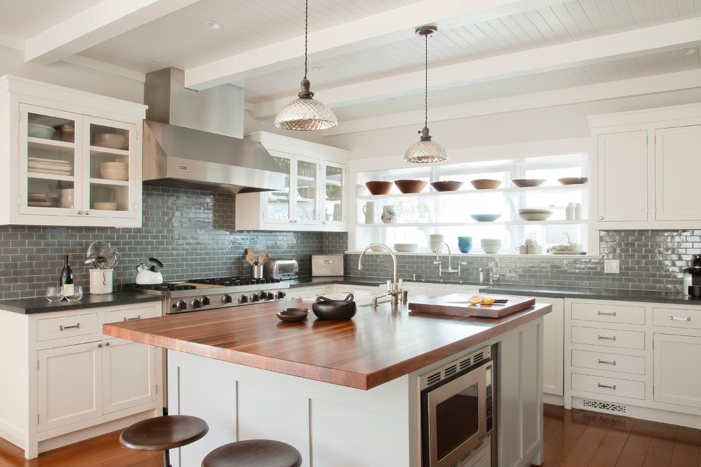 This is an example of a beach style kitchen in Los Angeles with glass-front cabinets, white cabinets, wood benchtops, grey splashback, subway tile splashback and stainless steel appliances.