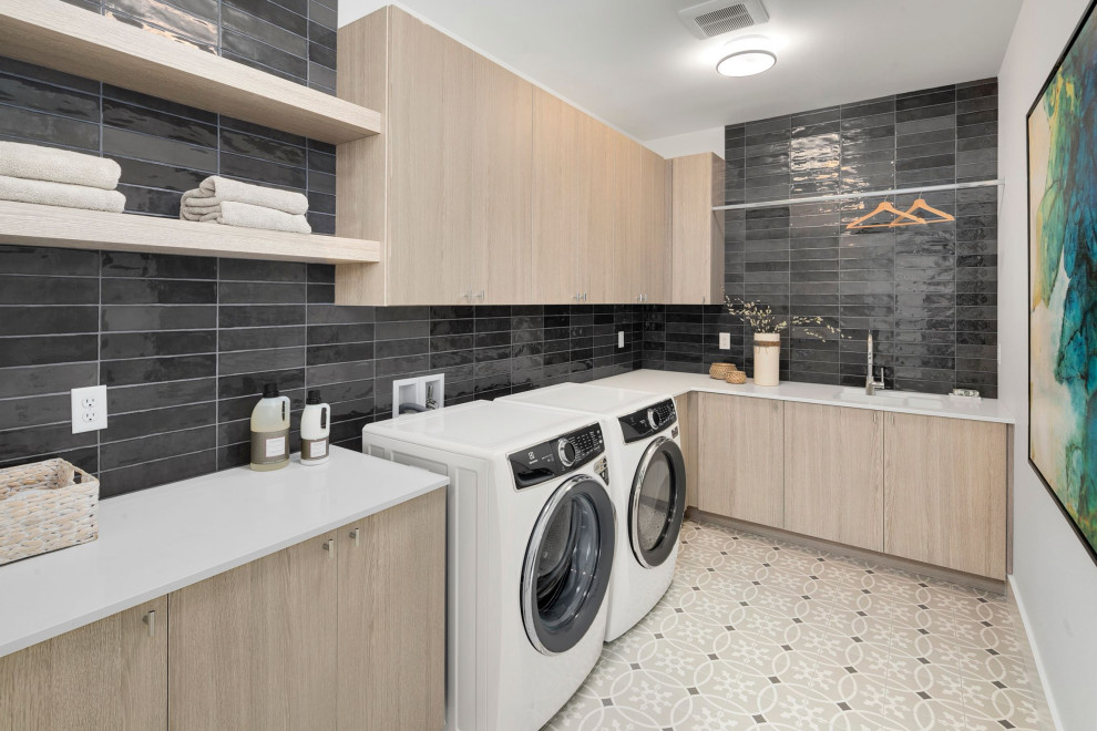 This is an example of a mid-sized l-shaped dedicated laundry room in Seattle with an utility sink, flat-panel cabinets, light wood cabinets, quartz benchtops, black splashback, subway tile splashback, white walls, porcelain floors, a side-by-side washer and dryer, beige floor and white benchtop.