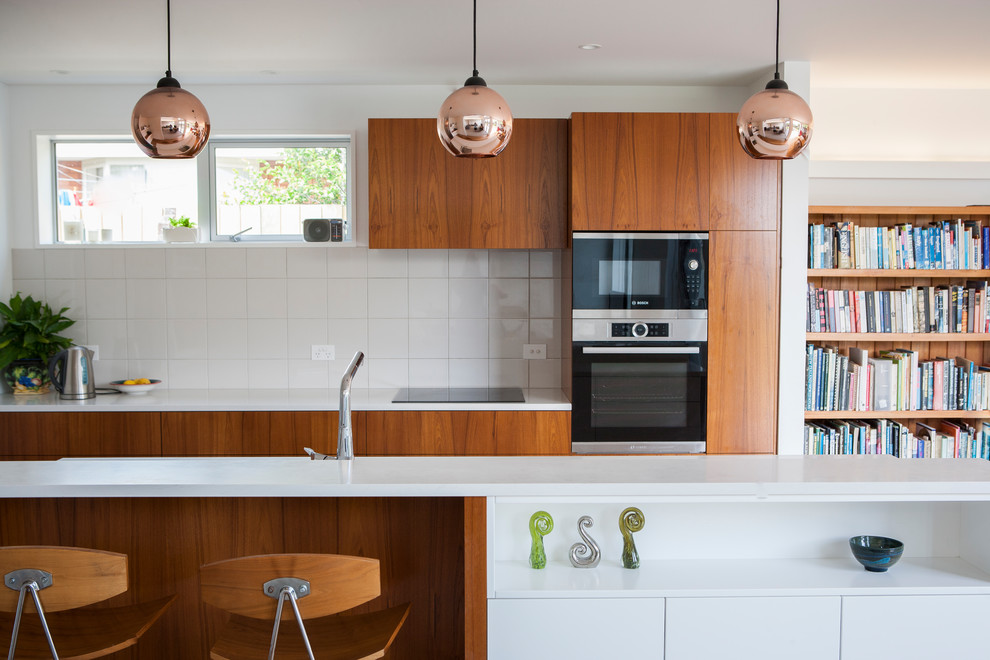 Photo of a contemporary galley open plan kitchen in Christchurch with flat-panel cabinets, medium wood cabinets, white splashback, black appliances, with island and white benchtop.