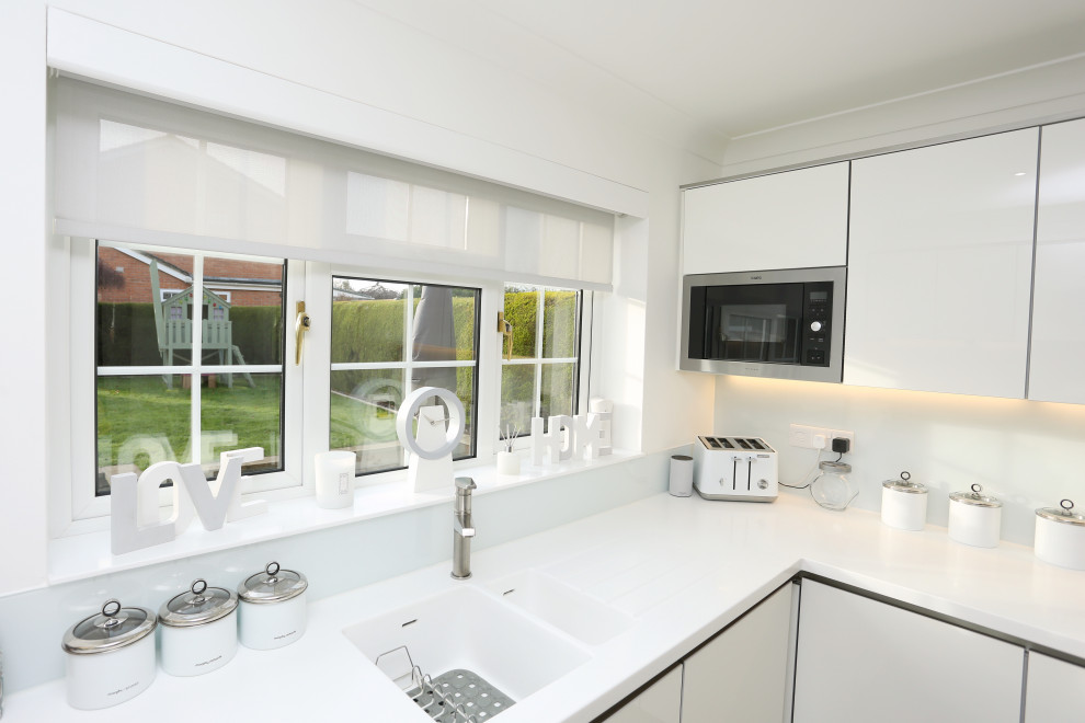This is an example of a mid-sized contemporary u-shaped separate kitchen in Other with an integrated sink, flat-panel cabinets, white cabinets, solid surface benchtops, black appliances, laminate floors, no island, brown floor and white benchtop.