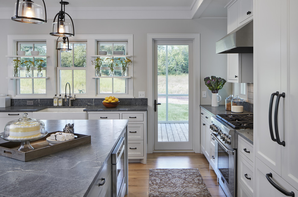 Mid-sized country l-shaped eat-in kitchen in Minneapolis with a farmhouse sink, white cabinets, soapstone benchtops, white splashback, light hardwood floors, with island, beaded inset cabinets, stone tile splashback and panelled appliances.