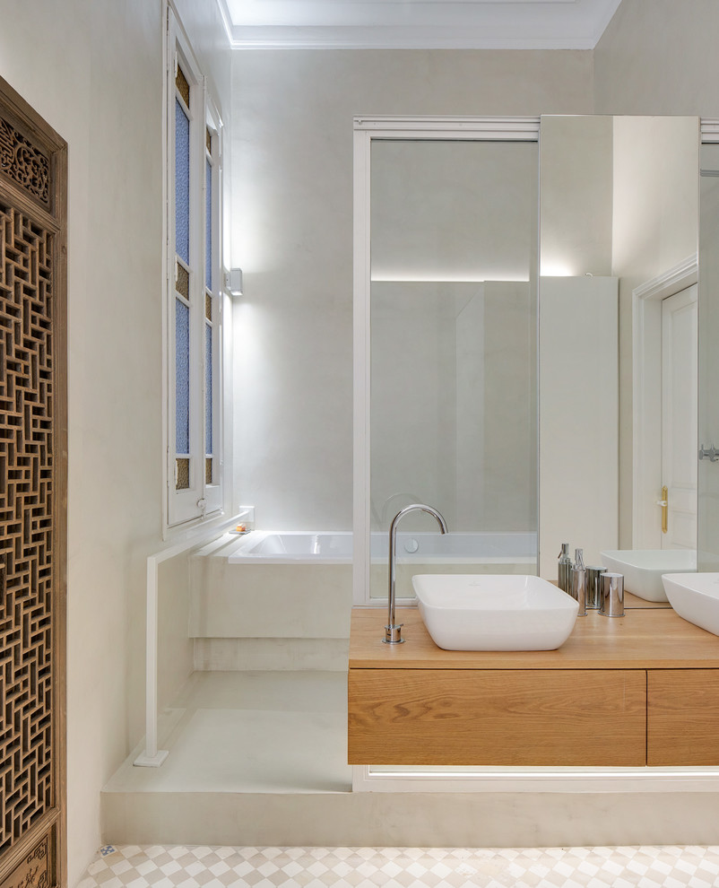 This is an example of a mid-sized mediterranean bathroom in Barcelona with an alcove tub, a vessel sink, wood benchtops, flat-panel cabinets, light wood cabinets, grey walls, grey floor and beige benchtops.
