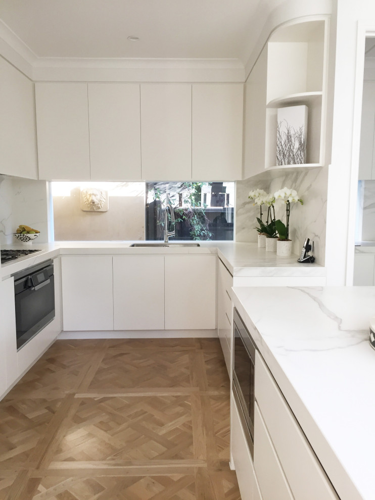 Mid-sized transitional u-shaped eat-in kitchen in Melbourne with a single-bowl sink, flat-panel cabinets, white cabinets, marble benchtops, white splashback, marble splashback, black appliances, light hardwood floors, with island, brown floor and white benchtop.