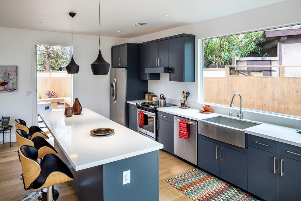 This is an example of a mid-sized contemporary single-wall kitchen in Los Angeles with flat-panel cabinets, blue cabinets, quartzite benchtops, stainless steel appliances, medium hardwood floors, with island, white benchtop, a farmhouse sink and brown floor.