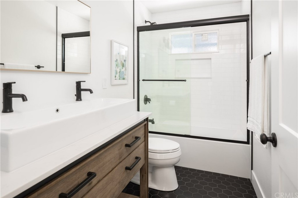 This is an example of a mid-sized country kids bathroom in Orange County with furniture-like cabinets, medium wood cabinets, an alcove tub, an alcove shower, a two-piece toilet, white tile, ceramic tile, white walls, ceramic floors, a vessel sink, engineered quartz benchtops, black floor, a sliding shower screen, white benchtops, a double vanity and a freestanding vanity.