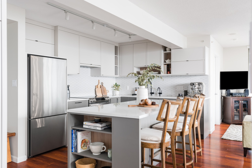 Inspiration for a mid-sized contemporary l-shaped eat-in kitchen in Vancouver with an undermount sink, shaker cabinets, white cabinets, quartz benchtops, white splashback, porcelain splashback, stainless steel appliances, medium hardwood floors, with island, brown floor and white benchtop.