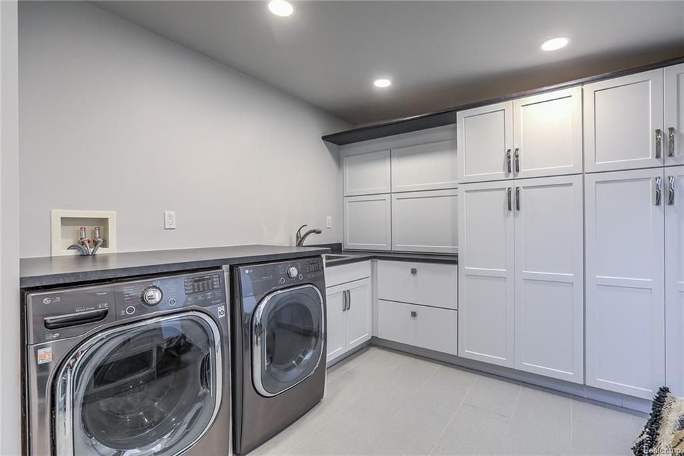 Design ideas for a mid-sized contemporary u-shaped utility room in Detroit with a drop-in sink, shaker cabinets, white cabinets, laminate benchtops, grey walls, porcelain floors, a side-by-side washer and dryer, grey floor and black benchtop.