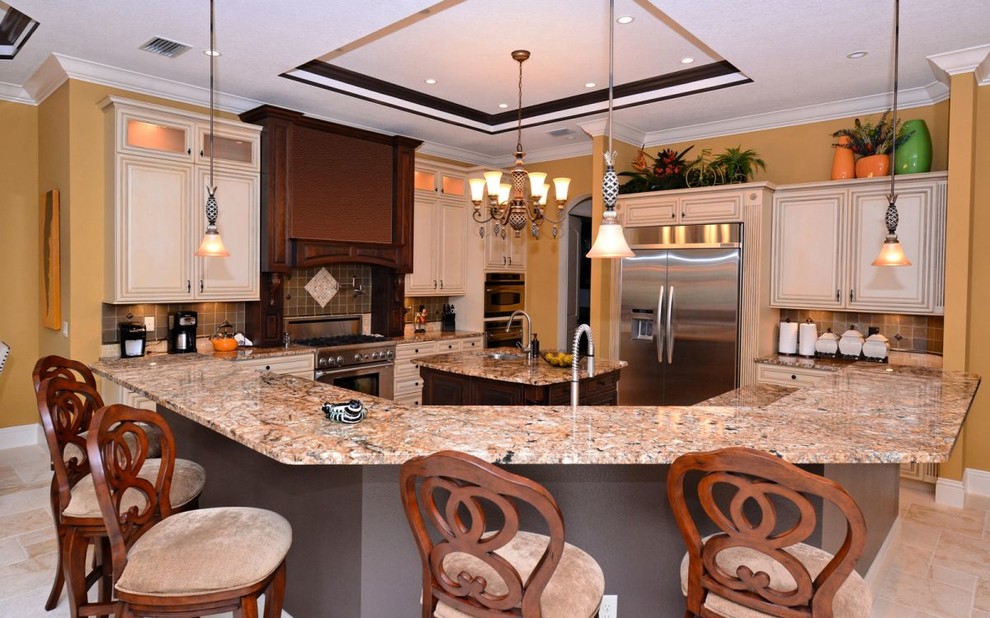 This is an example of a large mediterranean l-shaped open plan kitchen in Tampa with a drop-in sink, raised-panel cabinets, dark wood cabinets, granite benchtops, stainless steel appliances, limestone floors and multiple islands.