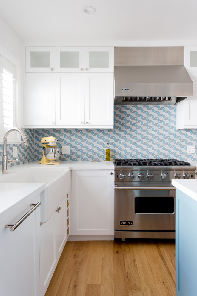 Example of a small beach style l-shaped light wood floor and brown floor enclosed kitchen design in Los Angeles with a farmhouse sink, shaker cabinets, white cabinets, solid surface countertops, blue backsplash, ceramic backsplash, stainless steel appliances, an island and white countertops
