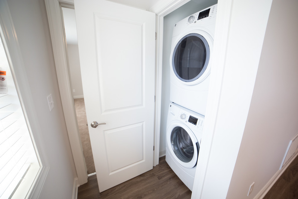 Photo of a small transitional laundry cupboard in New York with white walls, dark hardwood floors, a stacked washer and dryer and brown floor.
