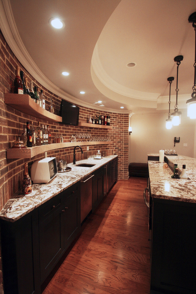 Photo of a large modern galley seated home bar in New York with an undermount sink, recessed-panel cabinets, dark wood cabinets, marble benchtops, red splashback, brick splashback, medium hardwood floors and brown floor.