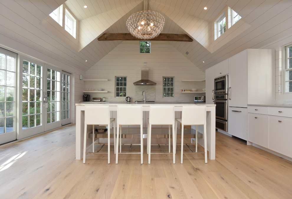 This is an example of a mid-sized beach style l-shaped open plan kitchen in Boston with flat-panel cabinets, white cabinets, white splashback, timber splashback, stainless steel appliances, light hardwood floors, with island and beige floor.