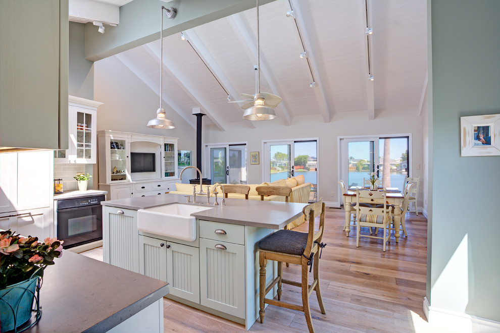 Photo of a mid-sized beach style l-shaped open plan kitchen in San Francisco with a farmhouse sink, shaker cabinets, white cabinets, quartz benchtops, blue splashback, subway tile splashback, panelled appliances, light hardwood floors and with island.
