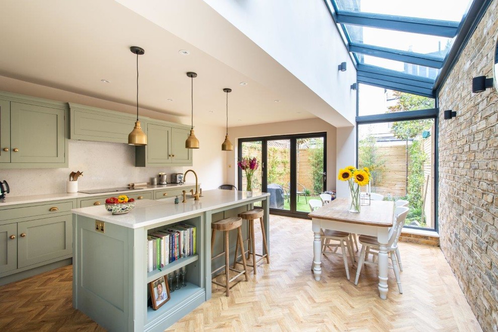 This is an example of a mid-sized transitional single-wall eat-in kitchen in Kent with green cabinets, white splashback, light hardwood floors, with island, beige floor, white benchtop and shaker cabinets.