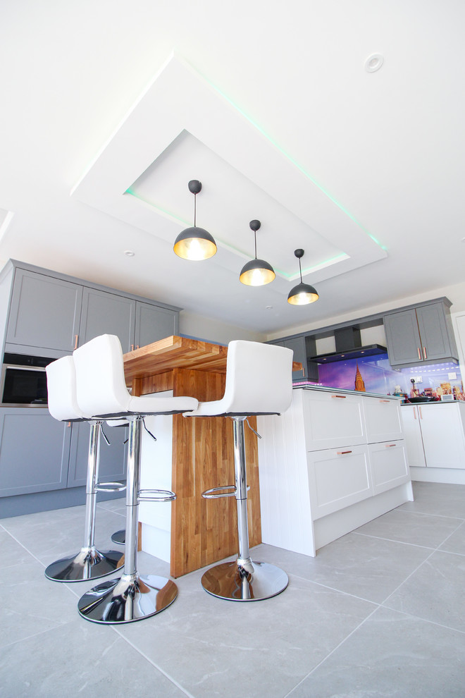 Photo of an expansive contemporary u-shaped open plan kitchen in London with an integrated sink, shaker cabinets, white cabinets, glass benchtops, multi-coloured splashback, glass sheet splashback, black appliances, ceramic floors, with island, grey floor and green benchtop.