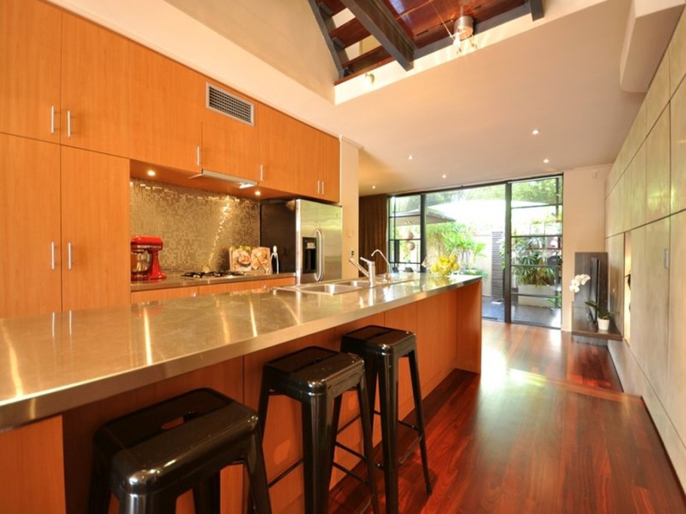 Photo of a small galley kitchen in Perth with an integrated sink, recessed-panel cabinets, light wood cabinets, stainless steel benchtops, metallic splashback, metal splashback, stainless steel appliances, dark hardwood floors and with island.
