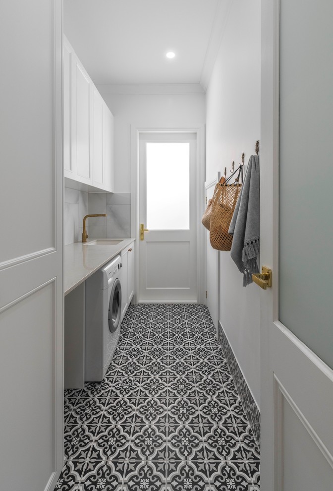 This is an example of a large beach style single-wall dedicated laundry room with an undermount sink, white cabinets, white walls, ceramic floors, a side-by-side washer and dryer, black floor and white benchtop.