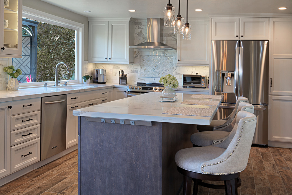 Photo of a mid-sized eclectic l-shaped open plan kitchen in Orange County with an undermount sink, recessed-panel cabinets, white cabinets, quartz benchtops, grey splashback, marble splashback, stainless steel appliances, porcelain floors, with island, brown floor and white benchtop.