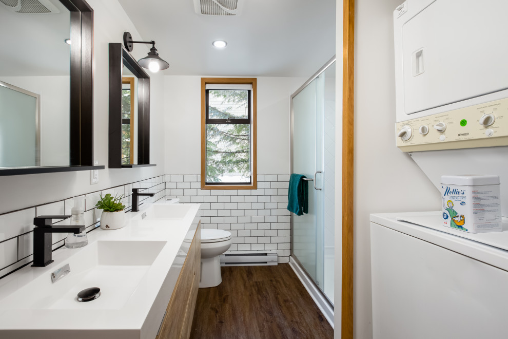 This is an example of a modern bathroom in Vancouver with flat-panel cabinets, light wood cabinets, a two-piece toilet, white tile, medium hardwood floors, brown floor, a sliding shower screen, a laundry, a double vanity, a freestanding vanity, a shower/bathtub combo, porcelain tile, white walls, an integrated sink, laminate benchtops and white benchtops.