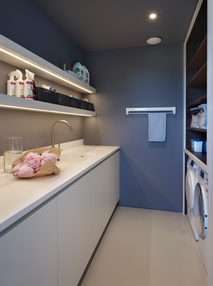 Photo of a mid-sized contemporary galley utility room in London with a double-bowl sink, flat-panel cabinets, white cabinets, solid surface benchtops, blue walls, a side-by-side washer and dryer, beige floor and white benchtop.