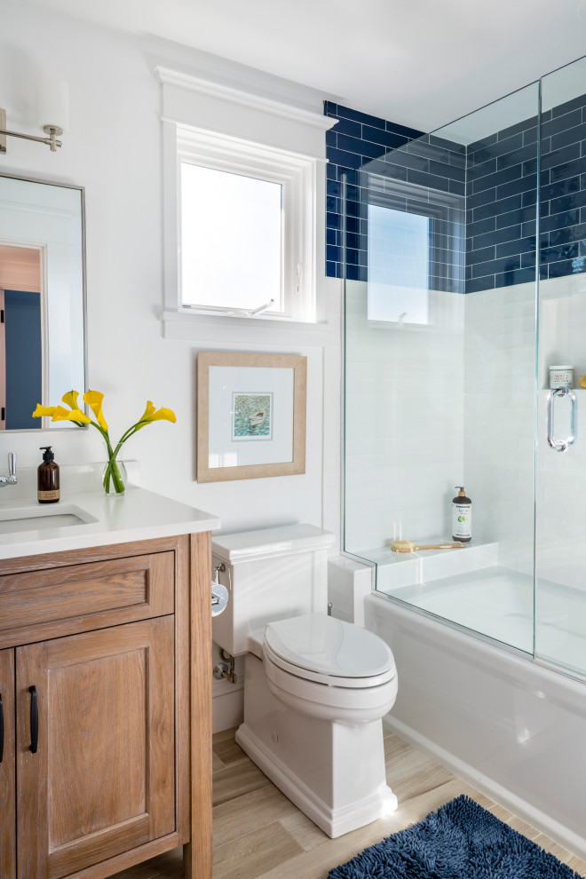 Photo of a beach style bathroom in Boston with shaker cabinets, medium wood cabinets, an alcove tub, a shower/bathtub combo, blue tile, white tile, white walls, an undermount sink, beige floor, a hinged shower door, white benchtops, a freestanding vanity and a single vanity.