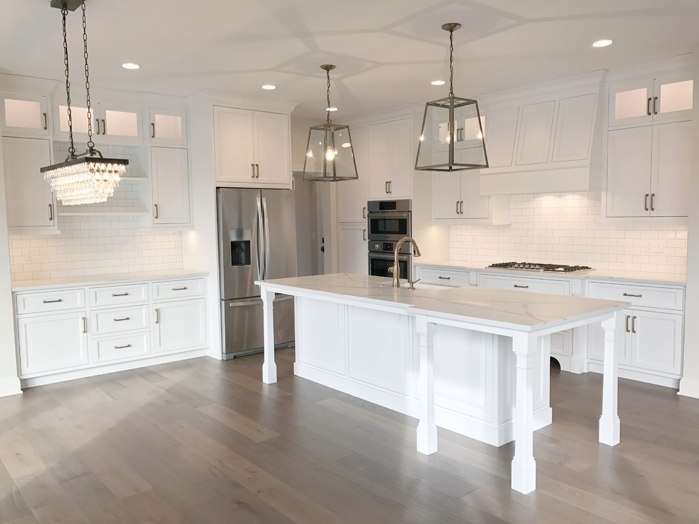 Inspiration for a large country l-shaped kitchen in Cleveland with an undermount sink, shaker cabinets, white cabinets, white splashback, subway tile splashback, stainless steel appliances, medium hardwood floors, with island and brown floor.