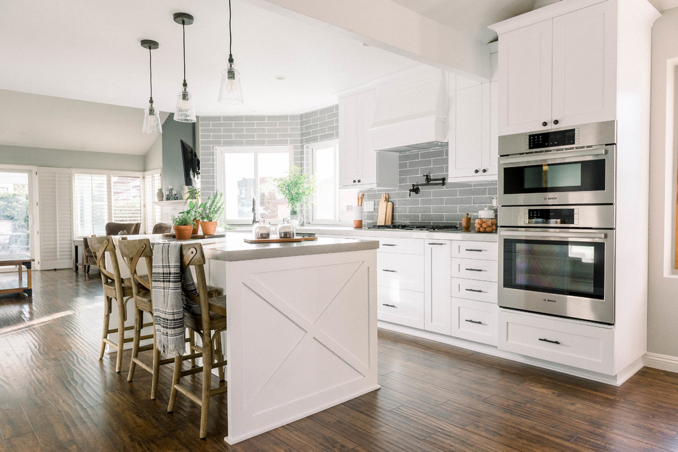 Photo of a mid-sized country single-wall open plan kitchen in San Diego with a farmhouse sink, shaker cabinets, white cabinets, quartz benchtops, grey splashback, ceramic splashback, stainless steel appliances, laminate floors, with island, brown floor and grey benchtop.