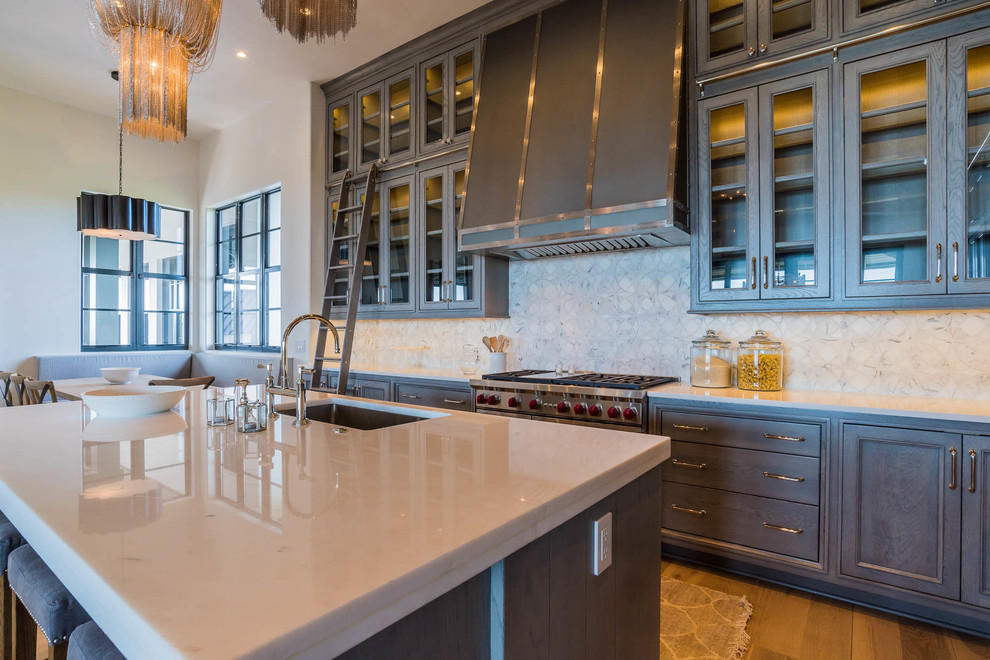 This is an example of a large beach style l-shaped open plan kitchen in Miami with an undermount sink, glass-front cabinets, grey cabinets, quartz benchtops, white splashback, stone slab splashback, stainless steel appliances, medium hardwood floors, with island, brown floor and grey benchtop.