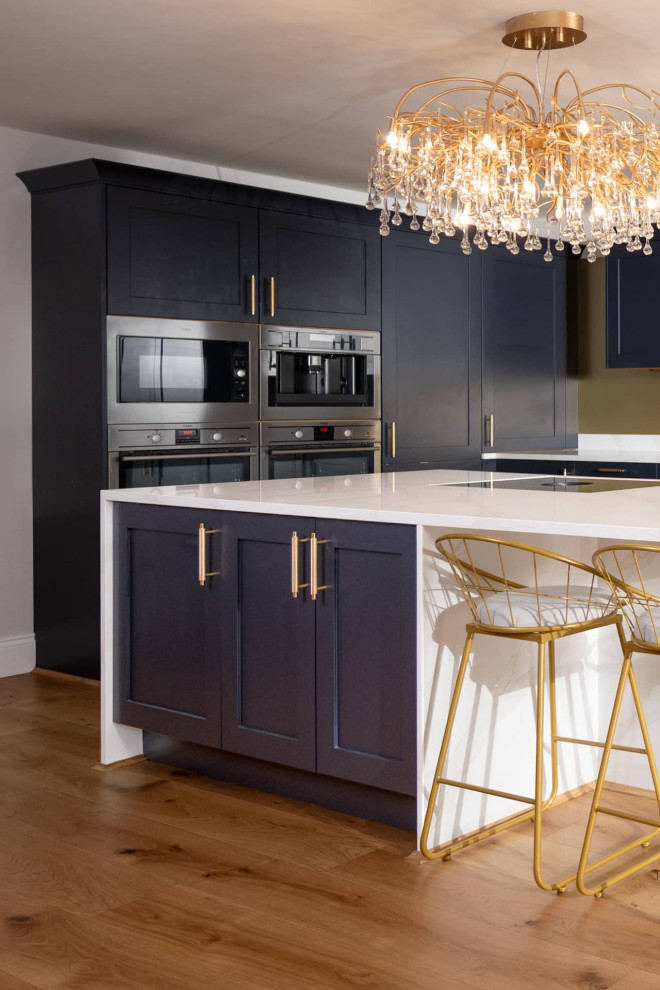 Inspiration for a large classic l-shaped open plan kitchen in Hertfordshire with a submerged sink, shaker cabinets, blue cabinets, quartz worktops, ceramic splashback, stainless steel appliances, light hardwood flooring, an island, brown floors, white worktops and feature lighting.