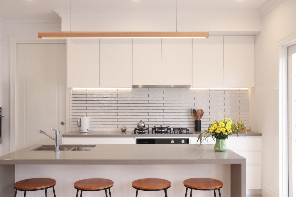 Photo of a large contemporary open plan kitchen in Melbourne with a double-bowl sink, flat-panel cabinets, white cabinets, quartz benchtops, white splashback, ceramic splashback, black appliances, light hardwood floors, with island, beige floor and grey benchtop.