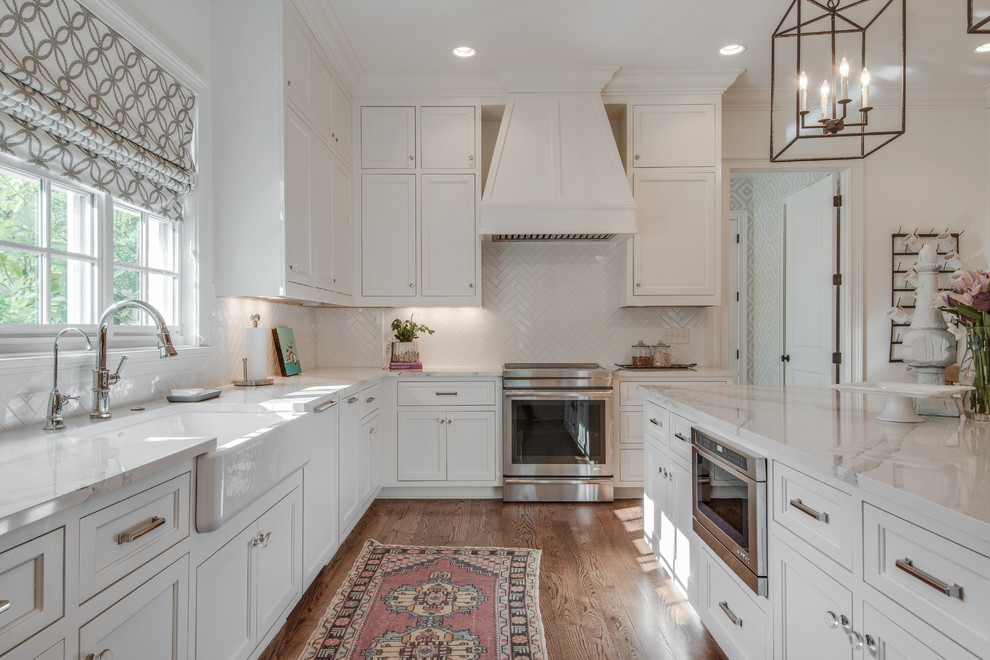 Inspiration for a large transitional l-shaped kitchen in Nashville with a farmhouse sink, recessed-panel cabinets, white cabinets, white splashback, with island, brown floor, quartz benchtops, porcelain splashback, panelled appliances and medium hardwood floors.