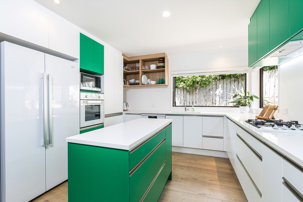 Photo of a mid-sized contemporary u-shaped eat-in kitchen in Auckland with an undermount sink, flat-panel cabinets, white cabinets, quartz benchtops, white splashback, glass sheet splashback, white appliances, with island, brown floor, medium hardwood floors and white benchtop.