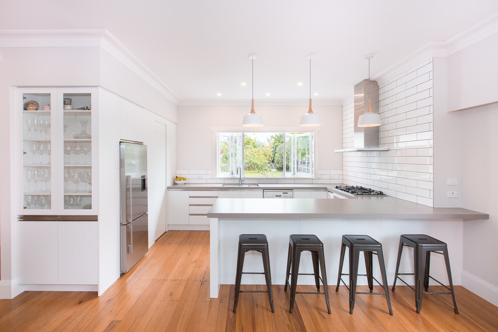 Photo of a contemporary u-shaped kitchen in Hamilton with an undermount sink, flat-panel cabinets, white cabinets, white splashback, subway tile splashback, stainless steel appliances, medium hardwood floors, a peninsula, brown floor and grey benchtop.