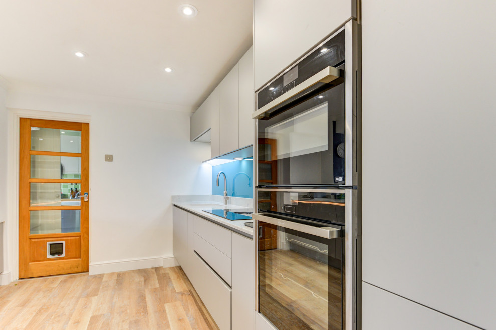 This is an example of a medium sized modern grey and white single-wall kitchen/diner in Sussex with a submerged sink, flat-panel cabinets, grey cabinets, quartz worktops, blue splashback, glass sheet splashback, integrated appliances, vinyl flooring, an island, brown floors, white worktops and feature lighting.