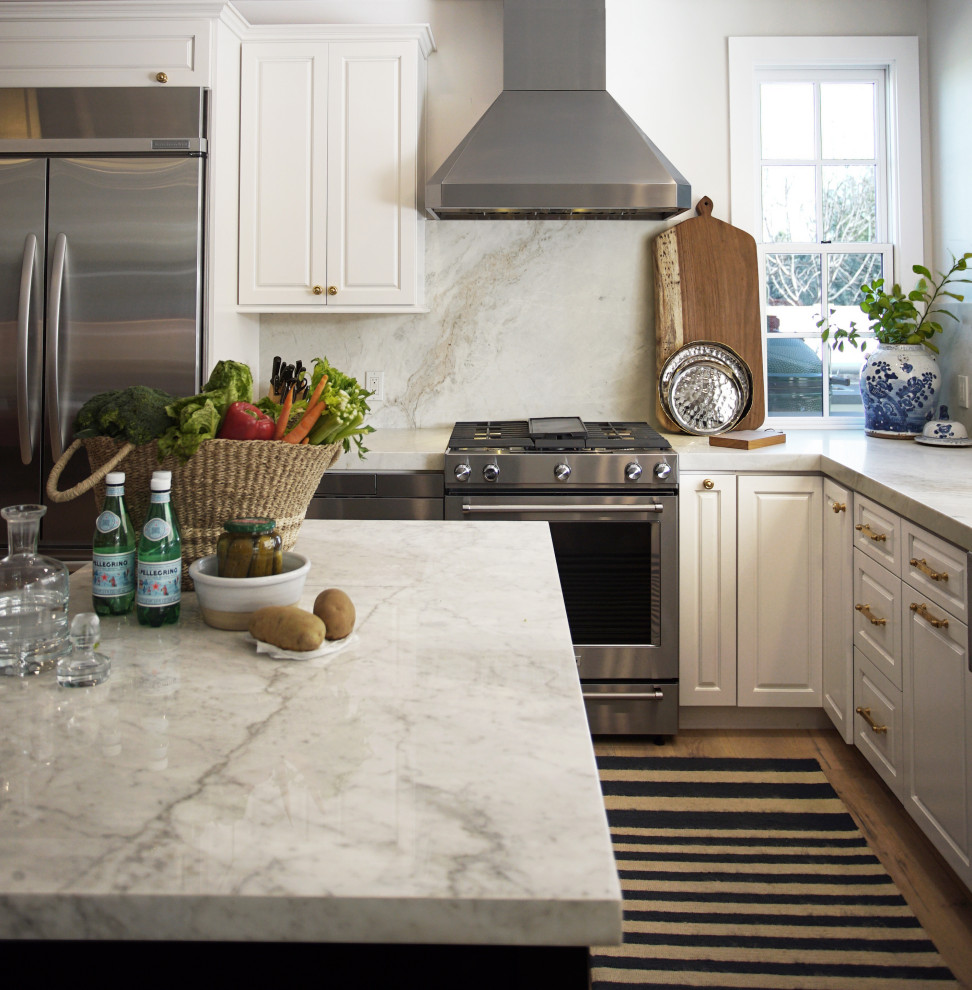 Mid-sized transitional l-shaped medium tone wood floor and brown floor open concept kitchen photo in Phoenix with a farmhouse sink, raised-panel cabinets, white cabinets, marble countertops, white backsplash, stone slab backsplash, stainless steel appliances, an island and white countertops