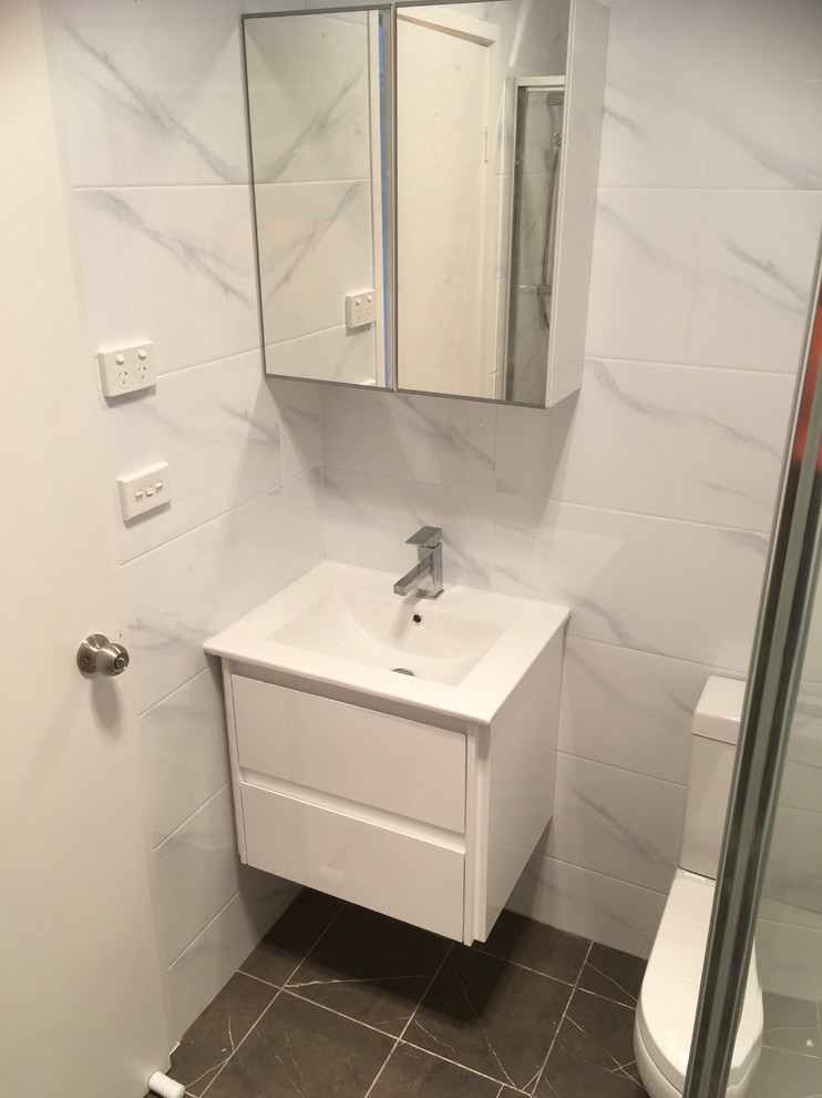 This is an example of a small contemporary 3/4 bathroom in Canberra - Queanbeyan with furniture-like cabinets, white cabinets, a corner shower, a one-piece toilet, white tile, ceramic tile, ceramic floors, a wall-mount sink, solid surface benchtops, grey floor, a hinged shower door and white benchtops.