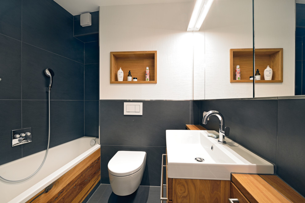 Photo of a small contemporary 3/4 bathroom in Frankfurt with recessed-panel cabinets, medium wood cabinets, a drop-in tub, a two-piece toilet, black tile, stone tile, white walls, terrazzo floors, a vessel sink, wood benchtops and black floor.