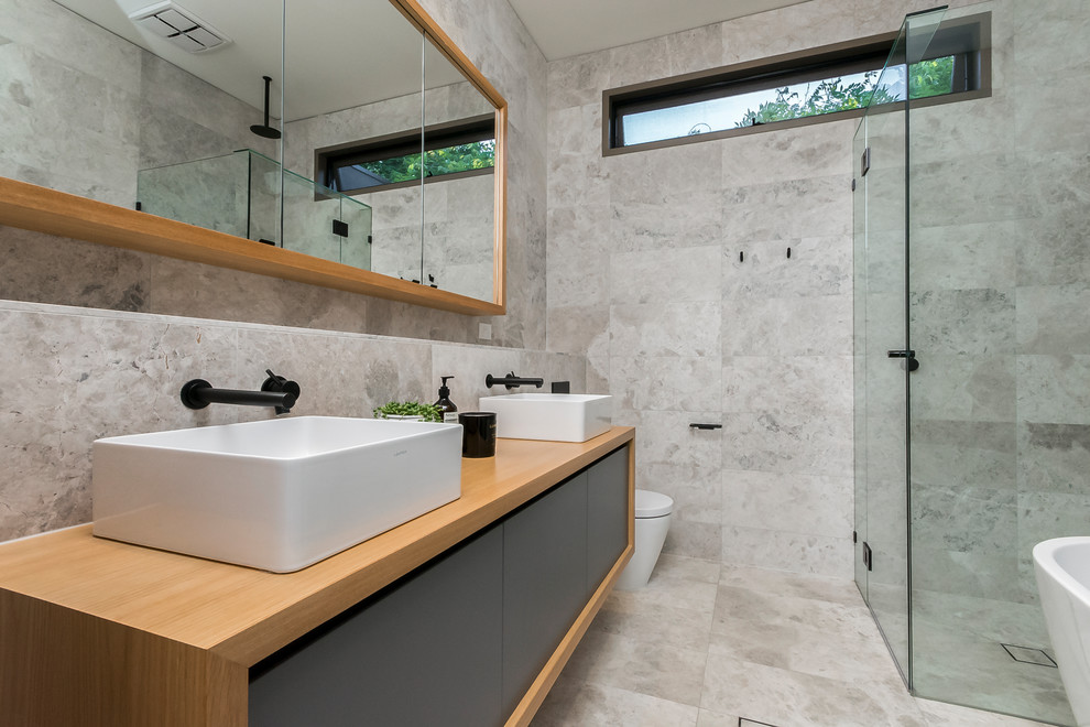 Design ideas for a contemporary master bathroom in Geelong with flat-panel cabinets, grey cabinets, a freestanding tub, a corner shower, a two-piece toilet, beige tile, gray tile, grey walls, a vessel sink, wood benchtops, grey floor and a hinged shower door.