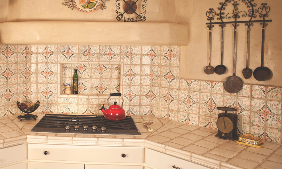 This is an example of a mid-sized mediterranean l-shaped eat-in kitchen in Albuquerque with a farmhouse sink, shaker cabinets, beige cabinets, tile benchtops, multi-coloured splashback, terra-cotta splashback, stainless steel appliances, terra-cotta floors and with island.