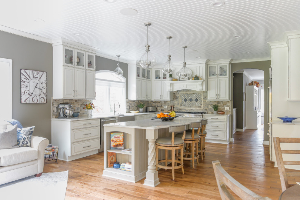 Inspiration for a large traditional l-shaped eat-in kitchen in Detroit with a farmhouse sink, raised-panel cabinets, white cabinets, quartz benchtops, beige splashback, travertine splashback, stainless steel appliances, medium hardwood floors, with island, brown floor and white benchtop.