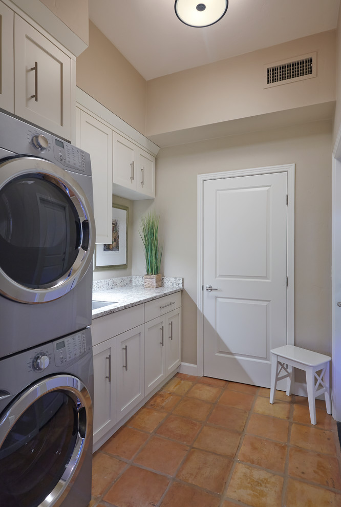 This is an example of a transitional single-wall laundry room in Phoenix with an undermount sink, shaker cabinets, white cabinets, granite benchtops, beige walls, terra-cotta floors and a stacked washer and dryer.