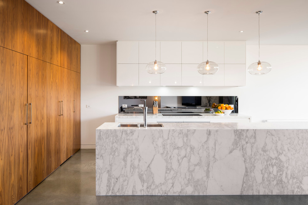 This is an example of a mid-sized contemporary l-shaped open plan kitchen in Melbourne with an undermount sink, flat-panel cabinets, white cabinets, marble benchtops, metallic splashback, glass sheet splashback, concrete floors and with island.