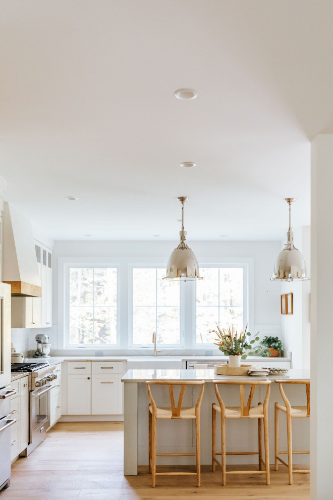 This is an example of a mid-sized transitional l-shaped kitchen in Portland Maine with a farmhouse sink, shaker cabinets, white cabinets, white splashback, subway tile splashback, stainless steel appliances, light hardwood floors, with island, beige floor and white benchtop.