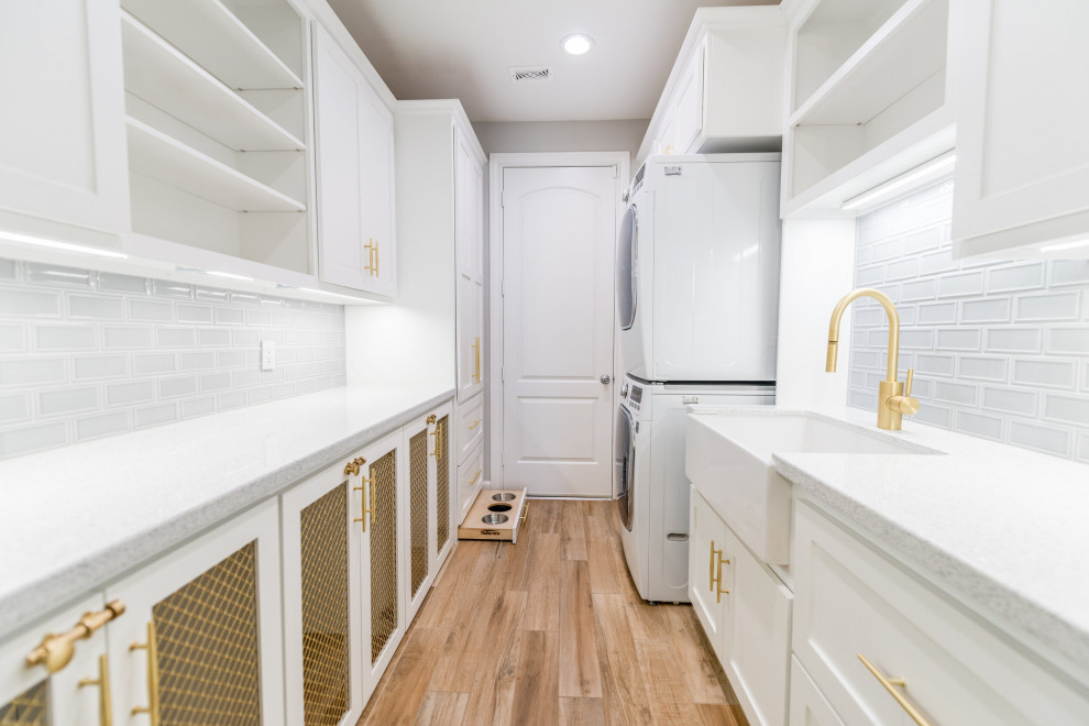 Inspiration for a large modern galley dedicated laundry room in Houston with a farmhouse sink, shaker cabinets, white cabinets, quartzite benchtops, grey splashback, subway tile splashback, grey walls, vinyl floors, a stacked washer and dryer, brown floor and white benchtop.