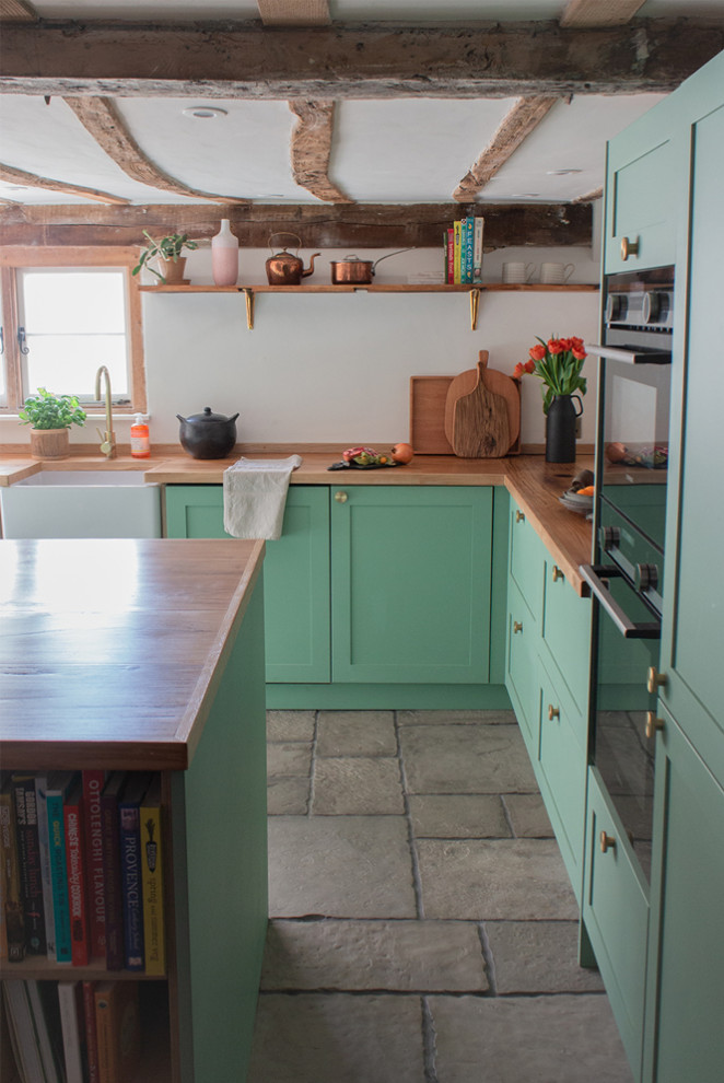 Photo of a mid-sized contemporary u-shaped eat-in kitchen in Oxfordshire with a farmhouse sink, shaker cabinets, green cabinets, wood benchtops, stainless steel appliances, limestone floors, with island, grey floor, brown benchtop and exposed beam.