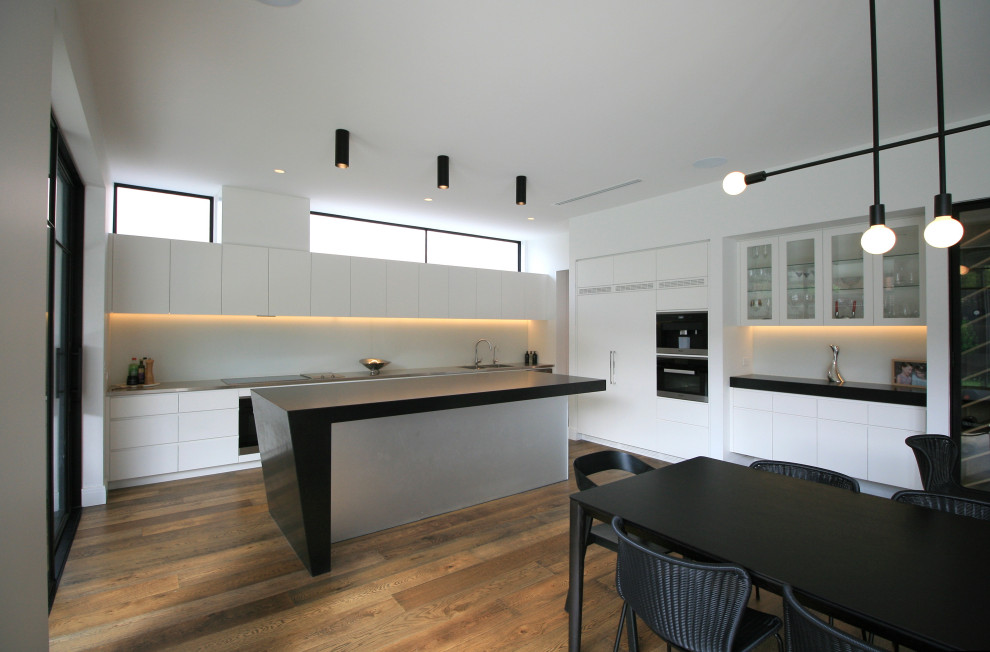 Large contemporary l-shaped open plan kitchen in Melbourne with a double-bowl sink, flat-panel cabinets, white cabinets, granite benchtops, white splashback, glass sheet splashback, black appliances, medium hardwood floors and with island.